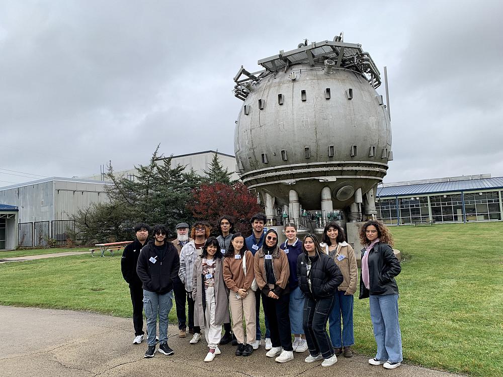 A group of people stand outdoors in front of a huge spherical steel chamber.